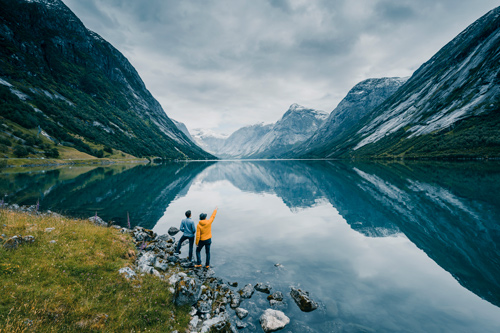 Två personer som står vid en sjö med stora berg i bakgrunden